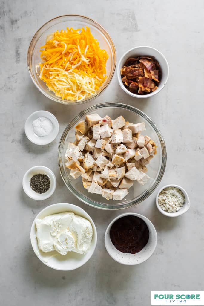 Aerial view of a clear glass bowl of cooked and cubed light meat chicken, a medium clear glass bowl of shredded white and yellow cheese and small white bowls of cooked chopped bacon, cream cheese, BBQ sauce, salt, pepper and ranch dressing mix. 