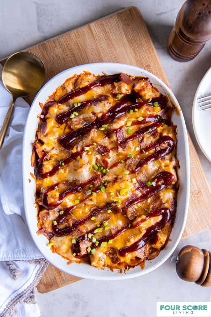 Aerial shot of white oval casserole dish with a baked BBQ chicken casserole drizzled with BBQ sauce and set on a light colored wood cutting board next to a white kitchen towel and a large brass round serving spoon all resting on a white natural stone surface. an edge of a white plate with a fork and wooden salt shaker and pepper grinder are in the right side background