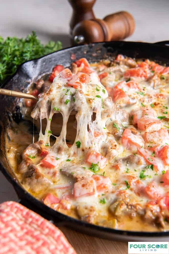 Diagonal view of a close up of Philly cheesesteak casserole in a cast iron pan with a serving being removed by a brass utensil. A red and white kitchen towel is in the foreground and a handful of fresh parsley and wooden salt and pepper grinders are in soft focus in the background.