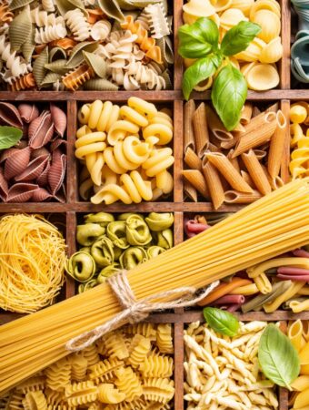 Aerial view of a wooden box with square compartments, each one filled with a different type of gluten free pasta of various colors and shapes.