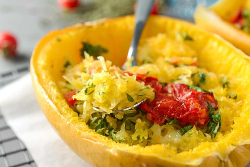Close up image of a front diagonal view of a halved, cooked spaghetti squash with red tomatoes and green herbs and a spoon lifting out a portion of the noodle-like squash flesh.