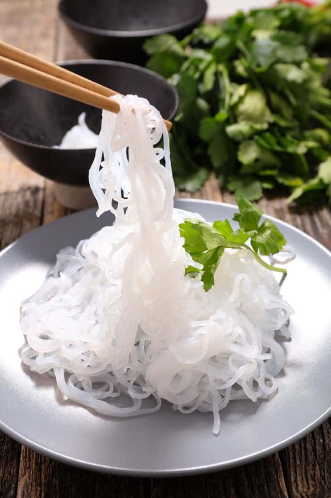 Diagonal view of shirataki noodles on a grey plate with chop sticks lifting a portion of noodles off of the plate, with a fresh green herb garnish on the noodles, a small black dish and fresh green herbs in soft focus in the background.
