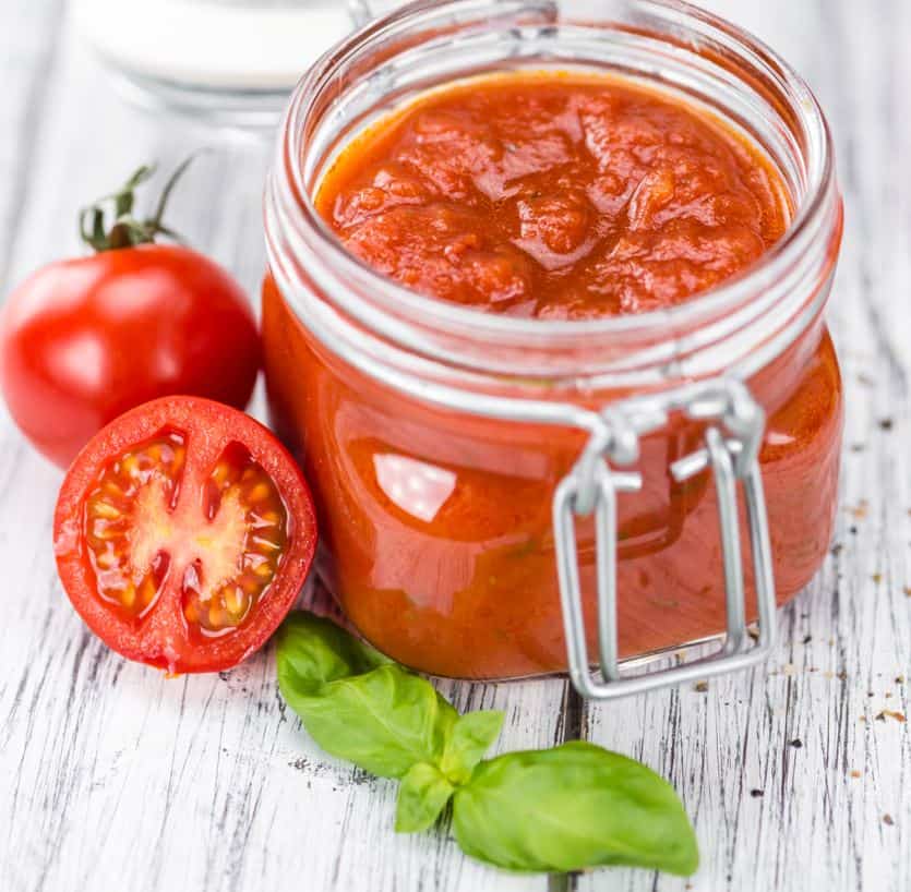 Glass mason jar with lid open filled with red tomato sauce, with one small, red vine tomato and one small vine red tomato cut in half with a spring of fresh basil all on a rustic whitewashed surface sprinkled with Italian seasoning. 