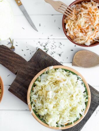 Aerial view of medium wooden bowls of sauerkraut, with on a dark wood cutting board and the remainder of items, including a sharp knife, wooden serving spoon, sprinkled dry herbs and a quarter of a fresh cabbage are all resting on a white bead board surface.