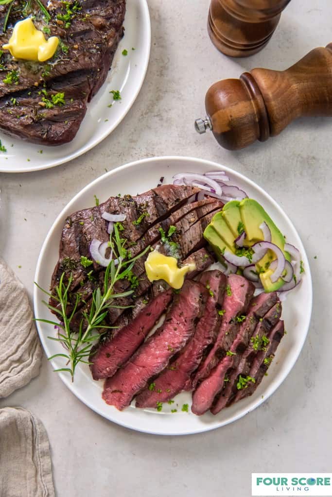 Diagonal, close up image of cooked 
 and sliced sirloin tip steak on a white dish with a pat of butter, fresh green herbs, a spring of rosemary, sliced ripe avocado and sliced red onion with wooden salt and pepper grinders and a whole, cooked sirloin steak in soft focus in the background.