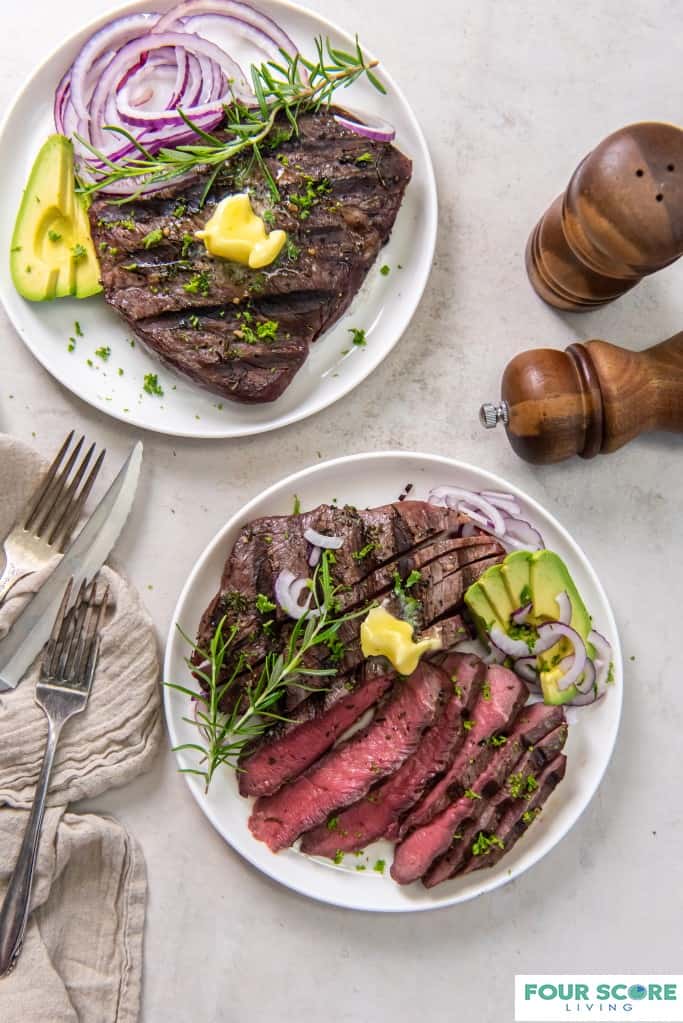 Aerial shot of two white plates of sirloin tip steak, one sliced and each garnished with sliced avocado, purple onion and a pat of butter. A kitchen towel, two forks and a steak knife, and wooden salt and pepper grinder all resting on a white stone surface. 