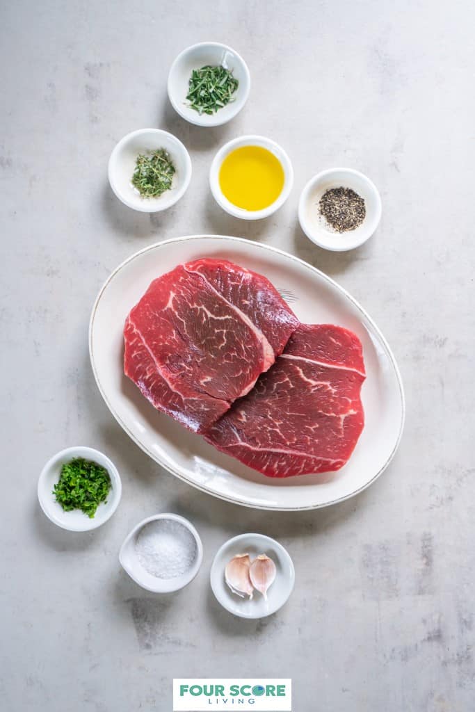 Aerial view of a white oval platter with two raw sirloin tip steaks and small dishes of herbs, spices, salt, two garlic cloves and olive oil. 