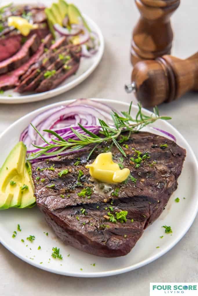 Diagonal, close up image of cooked sirloin tip steak on a white dish with a pat of butter, fresh green herbs, a spring of rosemary, sliced ripe avocado and sliced red onion with wooden salt and pepper grinders and a sliced sirloin steak in soft focus in the background. 