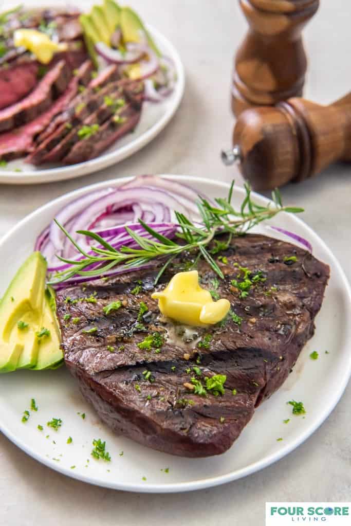Diagonal, close up image of cooked sirloin tip steak on a white dish with a pat of butter, fresh green herbs, a spring of rosemary, sliced ripe avocado and sliced red onion with wooden salt and pepper grinders and a sliced sirloin steak in soft focus in the background.