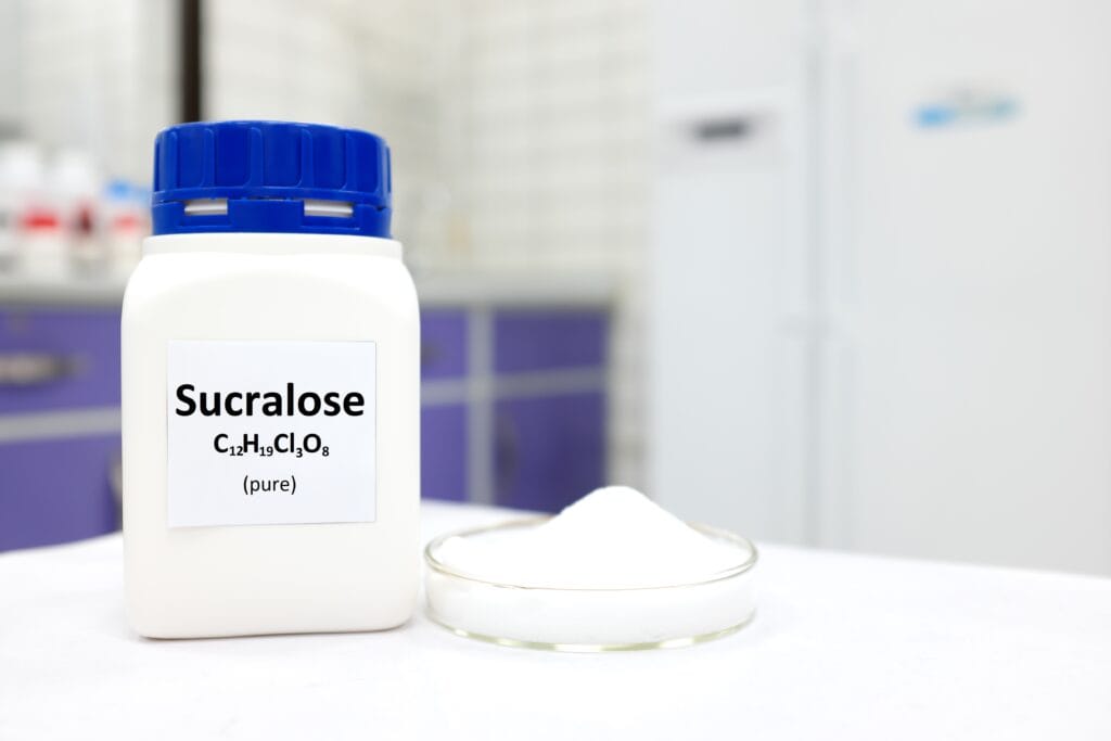 Large container of sucralose with a bright blue top in a laboratory setting with a white shallow dish of powered sucralose on a white surface. 