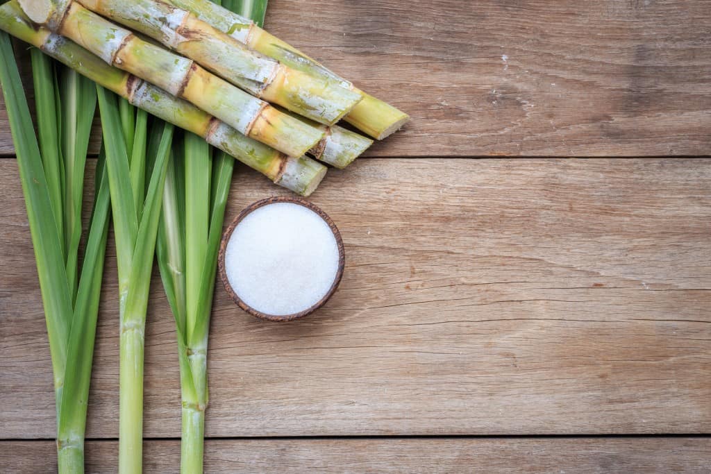 Sugarcane stalks and leaves on a rustic wooden background with a small brown bowl of white sweetener placed nearby.