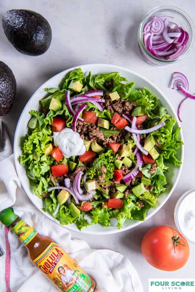 Taco salad in a white bowl with chopped tomatoes, sliced red onion, diced avocado, cooked ground beef and a dollop of sour cream with a white kitchen towel with a red strip, a whole red tomato, a bottle of hot sauce, a partial view of a dish of sour cream, whole avocados and a dish of sliced red onions, all on a white stone surface.