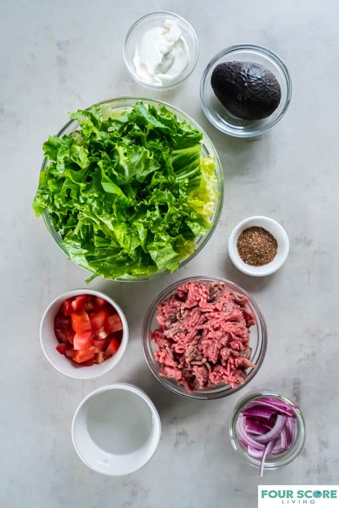 Ingredients to make a taco salad with chopped lettuce, raw ground beef, chopped tomatoes, taco seasoning, sour cream and a whole avocado in clear glass and white bowls.