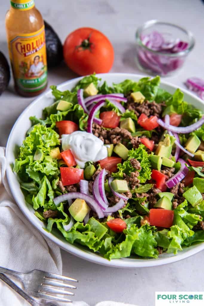 Close up diagonal view of taco salad in a white bowl with chopped tomatoes, sliced red onion, diced avocado, cooked ground beef and a dollop of sour cream with a white kitchen towel a fork, a whole red tomato, a bottle of hot sauce, a partial view of a dish of sour cream, whole avocados and a dish of sliced red onions, all on a white stone surface.