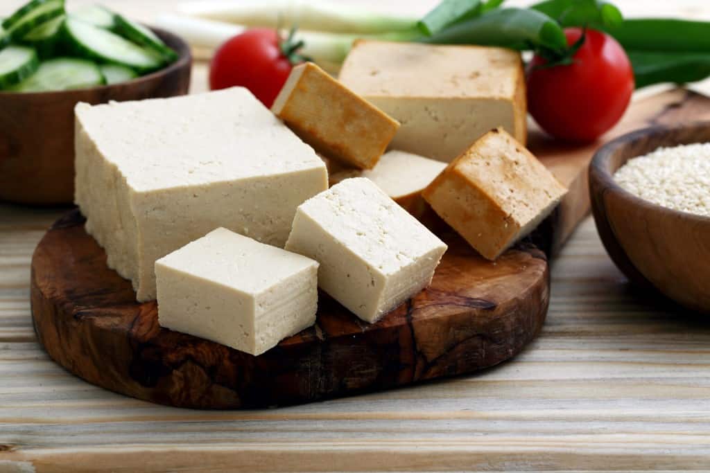 Diagonal view of blocks and cubes of tofu on a cutting board with two red small whole tomatoes and sliced cucumbers in a bowl in soft focus in the background.