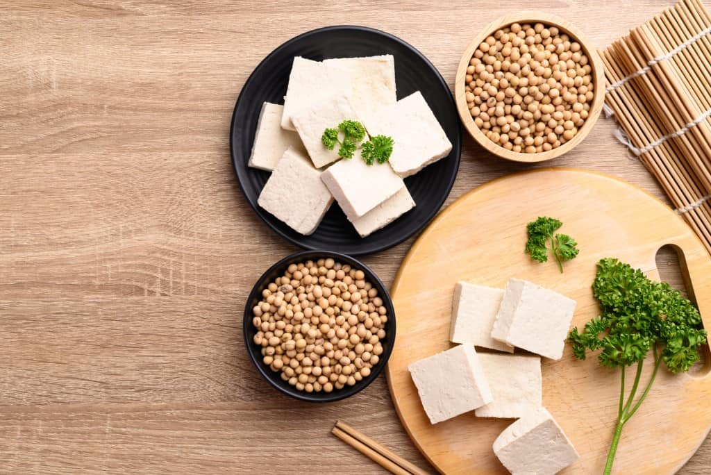 Aerial view of square blocks of tofu on a round light wood cutting board, a small black plate with fresh green garnish and two small bowls of soybeans on a wooden surface.