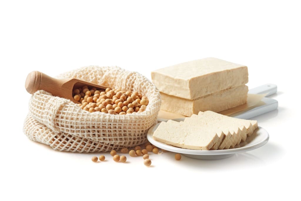 White cutting board with two blocks of tofu and white plate with sliced raw tofu and a natural sac rolled down overflowing with soybeans with a small wooden scoop all on a white background.
