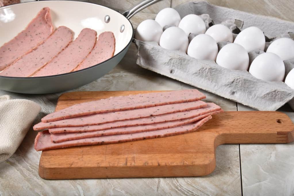 Uncooked slices of natural organic turkey bacon on a brown wooden cutting board with a ceramic fry pan layered with raw turkey bacon and a dozen of white eggs in an open egg carton in a front diagonal view.