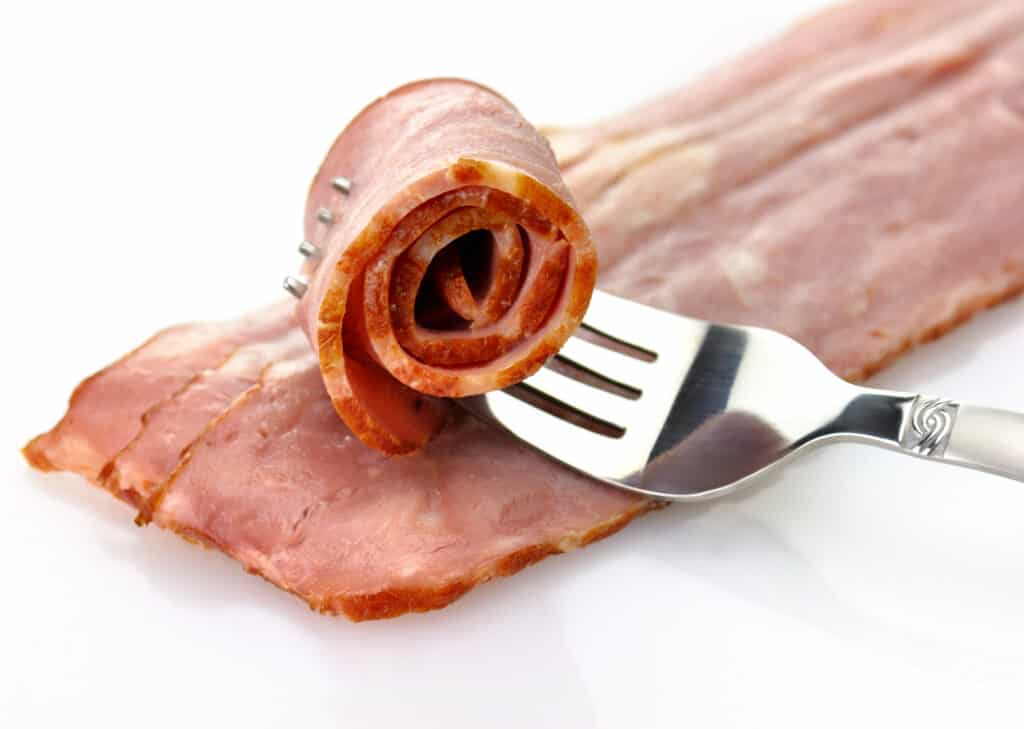 Diagonal, closeup view of three strips of raw turkey bacon with one rolled up strip of raw turkey bacon resting on top pierced by a fork all on a white background. 