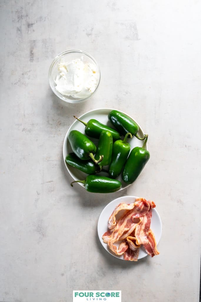 Aerial view of a white dish with 8 whole, fresh jalapenos, a dish of 8 strips of precooked bacon and a small glass dish of cream cheese.