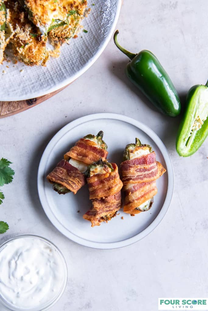 Aerial view of three bacon wrapped cream cheese filled jalapeno poppers on a small plate with a fresh whole and halved jalapeno, a small glass dish of cream sauce and a plate of air fryer jalapeno poppers in the perimeter of the image.