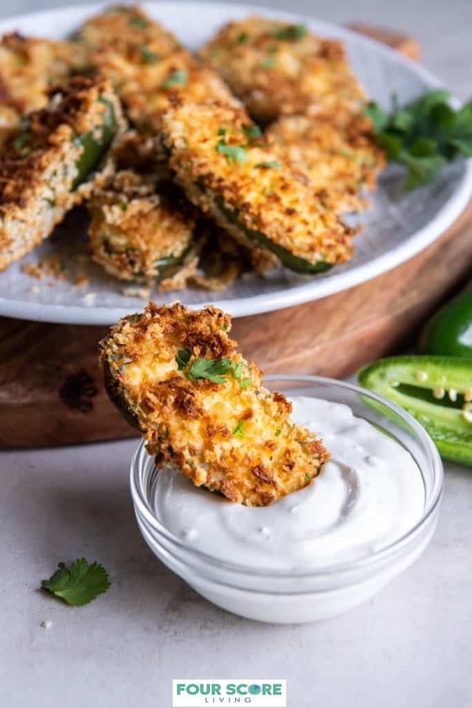 Frontal view of a prepared air fryer jalapeno popper with one end dipped into a small glass dish of white cream sauce with a halved fresh jalapeno and a cutting board with a white dish of air fryer jalapeno poppers in soft focus in the background. 