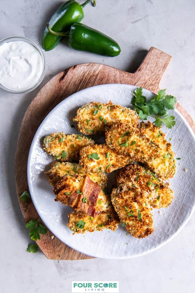 Aerial view of air fried jalapeno poppers on a white plate layered on top of a stone counter and a round cutting board, with one jalapeno popper wrapped in bacon, sprinkled with chopped fresh herbs.