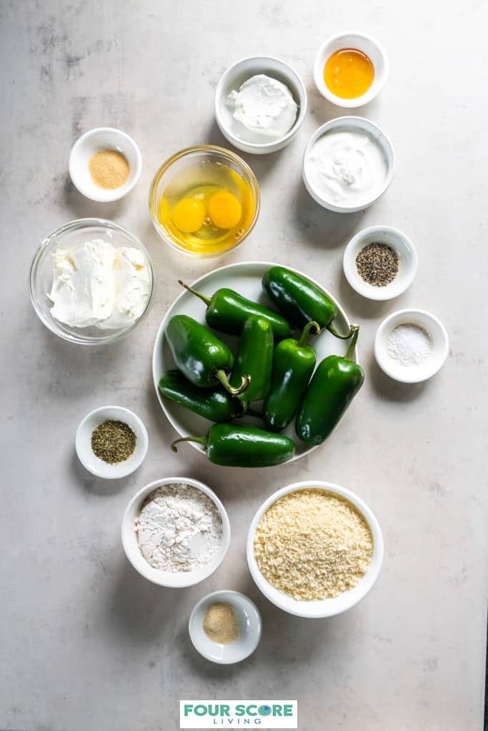 Aerial view of a ingredients to make air fryer jalapeno poppers, including a white dish of 8 whole, fresh jalapenos, dishes of salt and pepper, eggs, flour, cream cheese, bread crumbs and flour.  