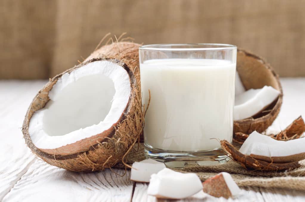 Front view of drinking glass of coconut milk on a natural hemp napkin on white wooden table with a fresh brown coconut broken into three pieces exposing the white flesh.