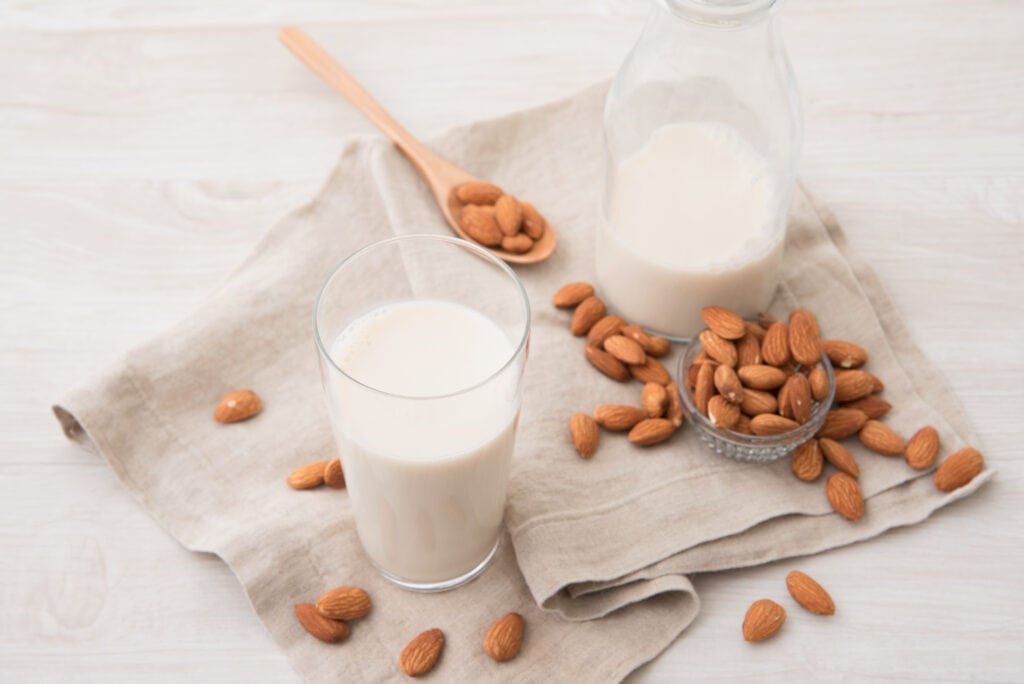 Aerial diagonal view of a small clear glass bottle one third of the way filled with almond milk, a glass more than half full of almond milk, a small glass dish overflowing with whole almonds, almonds spilled out onto the surface and a medium wooden spoon with almonds all resting on a natural fiber kitchen towel and a whitewashed wooden surface.