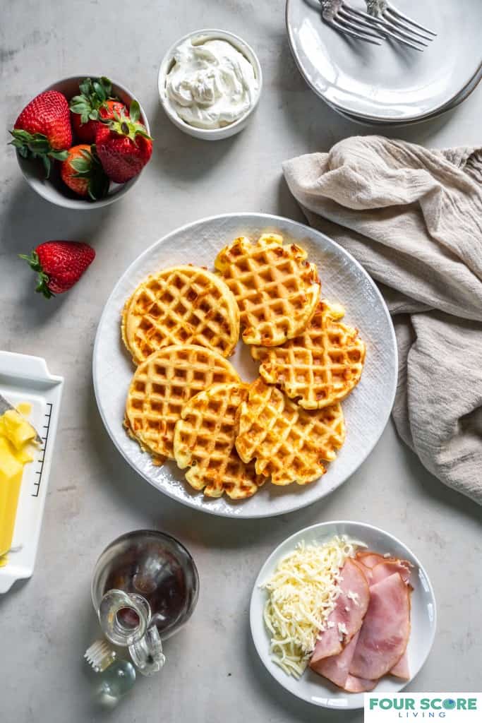 Aerial view of a white textured plate with six cooked Keto Chaffles surrounded on the nearby light stone surface by a stick of softened butter, a small plate with ham and shredded cheese, a glass bottle of sugar free syrup, a bowl of fresh strawberries, a small white dish of whipped cream, a natural colored kitchen towel, small white plates and forks.