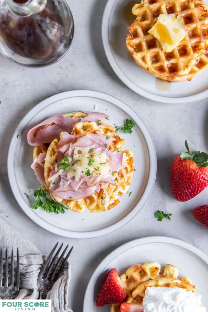Aerial view of a three cooked keto waffles, each on a small plate, one in prominent full view with fresh halved strawberries and whipped cream, one in full view with sliced ham, melted white cheese and green garnish, one in ¾ view with a pat of butter and sugar free syrup, with a glass bottle of syrup in soft focus to the left of the upper frame. 