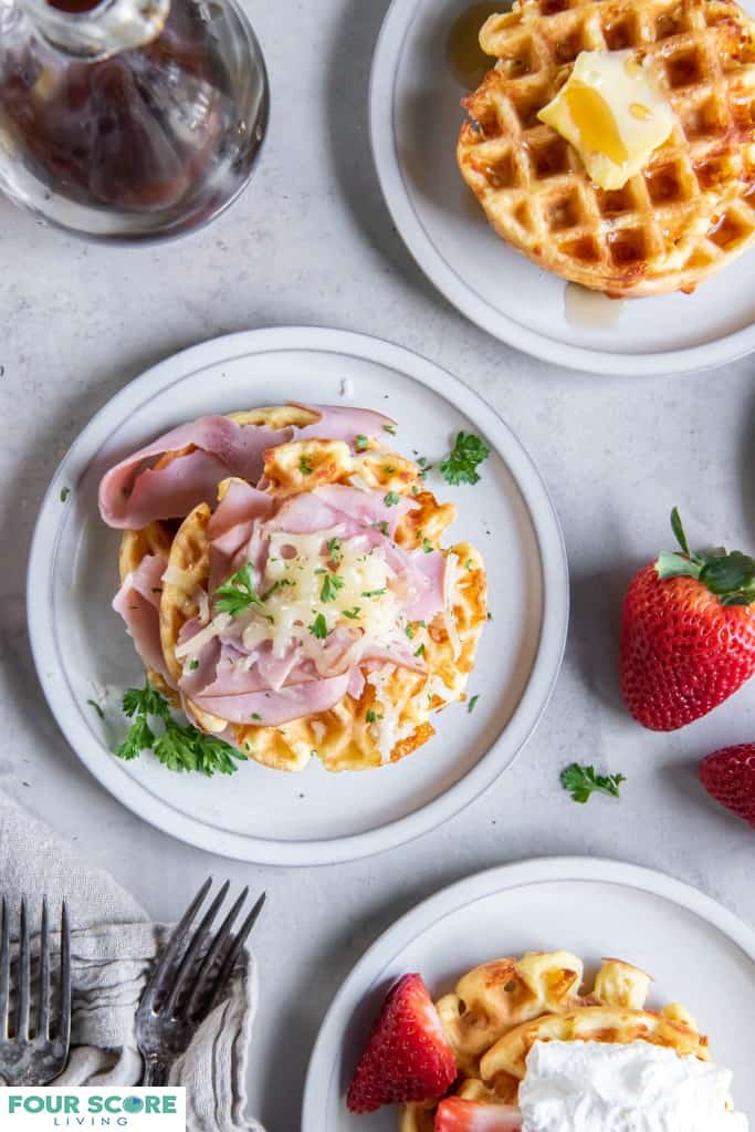 Aerial view of a three cooked keto waffles, each on a small plate, one in prominent full view with fresh halved strawberries and whipped cream, one in full view with sliced ham, melted white cheese and green garnish, one in ¾ view with a pat of butter and sugar free syrup, with a glass bottle of syrup in soft focus to the left of the upper frame.