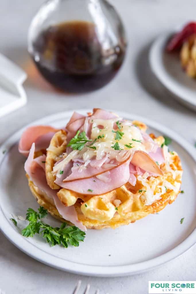 Diagonal aerial view of cooked Keto Chaffle in prominent full view with sliced ham, melted white cheese and green garnish on a white plate with a partial view of a bottle of maple syrup in soft focus in the background, all on a light stone surface.