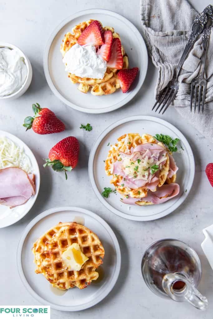Aerial view of a three cooked keto waffles, each on a small plate, all in prominent full view  one with fresh halved strawberries and whipped cream, one with sliced ham, melted white cheese and green garnish, one  with a pat of butter and sugar free syrup, with a glass bottle of syrup in the right lower corner of the frame, with three whole strawberries, a natural kitchen towel, two forks, a small dish of whipped cream and a partial view of a small plate of ham and shredded cheese all on a light colored stone surface. 