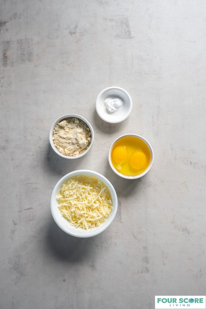 Aerial view of ingredients for making a Keto Chaffle, including a small to medium bowls containing raw eggs, a small dish of baking powder, a cup of shredded cheese, and a small dish of almond flour, all on a light colored stone surface.