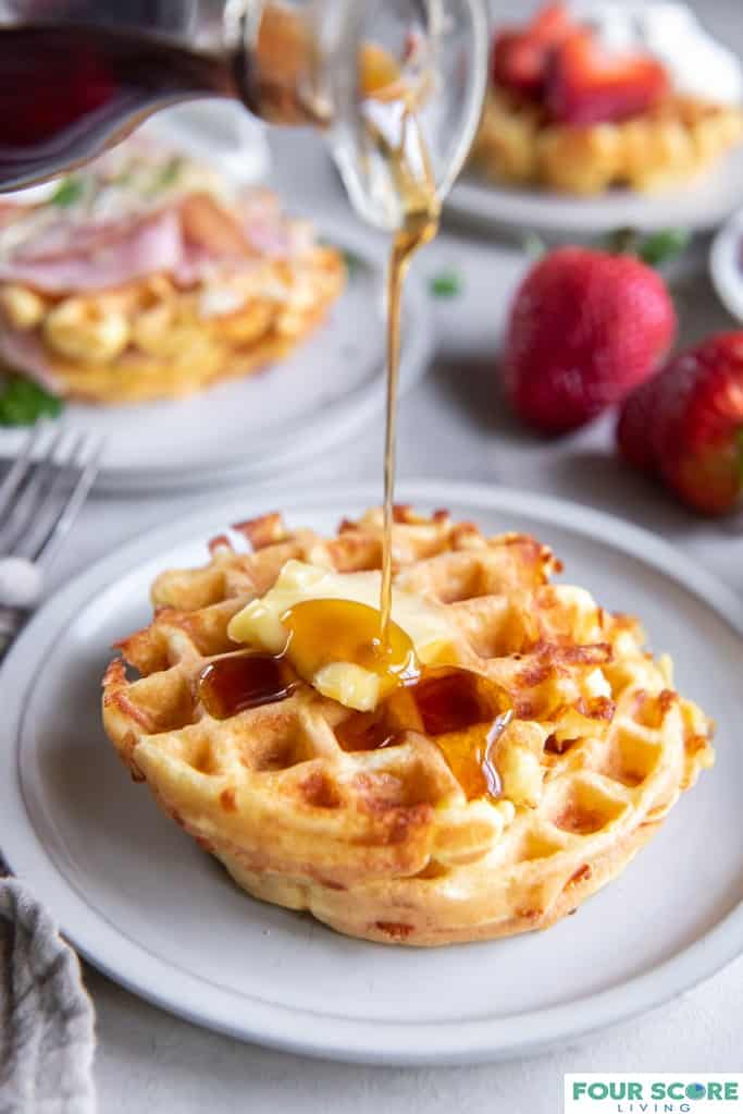 Aerial diagonal view of three cooked keto waffles, each on a small plate, one in ¾ view in soft focus in the background with fresh halved strawberries and whipped cream, one in full view with and in soft focus with sliced ham, melted white cheese and green garnish, one in full prominent view with a pat of butter and sugar free syrup pouring on top with two whole strawberries all resting on a light stone surface. 