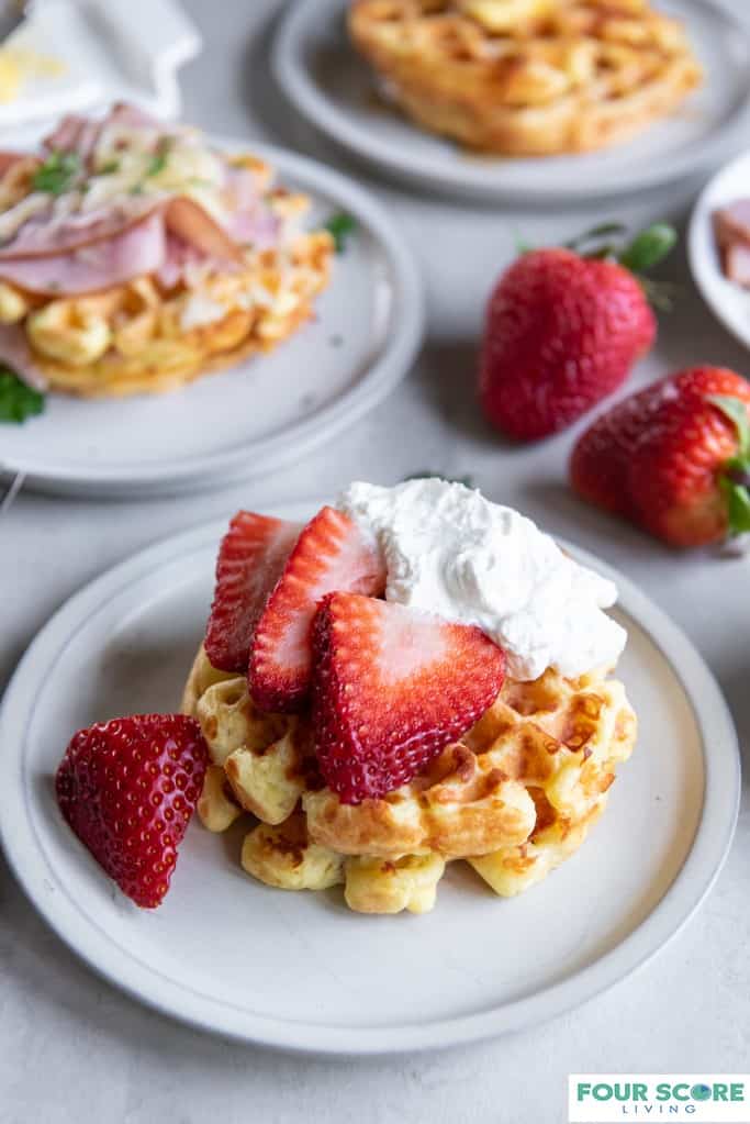 Aerial diagonal view of three cooked keto waffles, each on a small plate, one in full view with fresh halved strawberries and whipped cream, one in full view with  and in soft focus with sliced ham, melted white cheese and green garnish, one in ¾ view with a pat of butter and sugar free syrup, with two whole strawberries all resting on a light stone surface. 