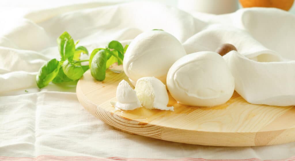 Three fresh balls of mozzarella cheese with two fresh sprigs of basil on a light colored, round, wood cutting board placed on top of a white kitchen towel with a red stripe. background.