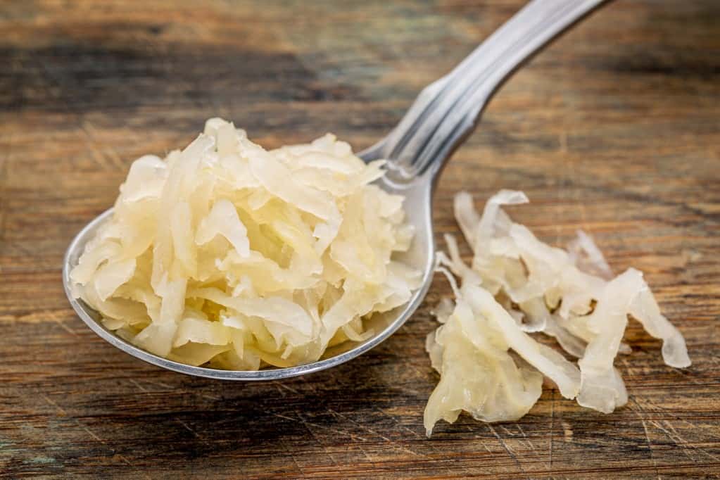 Close up diagonal view of a silver soup spoon filled with sauerkraut and a small amount of sauerkraut next to it on a rustic wooden surface.