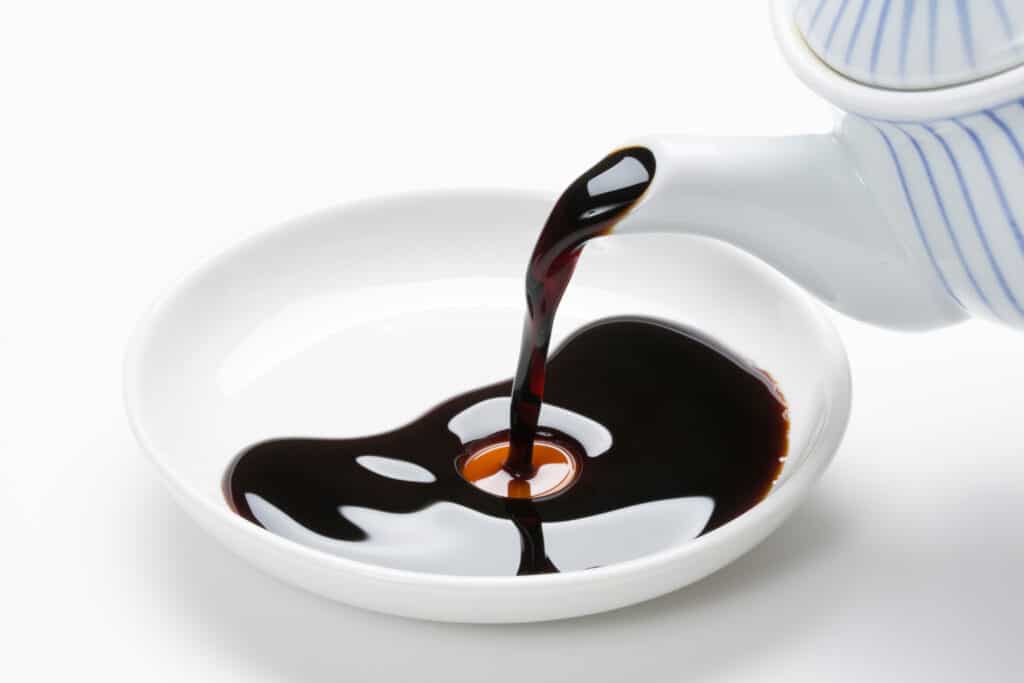 Frontal diagonal view of soy sauce being poured from a white ceramic tea pot with thin blue vertical stripes, into a white shallow dish on a white background. 