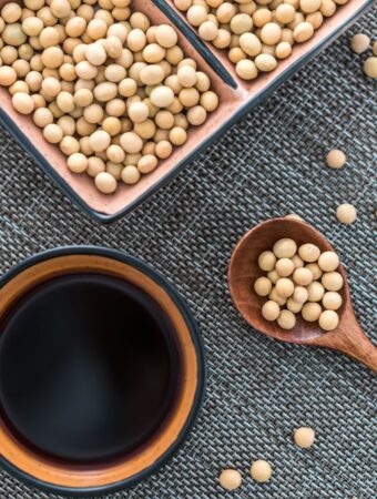 Aerial view of woven mat with a dish of soy sauce, a large wooden spoon and a rectangular dish filled with raw, whole soybeans spilling out onto the surface with a natural cloth kitchen towel in the upper left side of the frame.