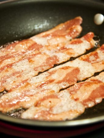 Frontal diagonal view of cooking slices turkey bacon in a fa dark colored frying pan. Dark surface in background.