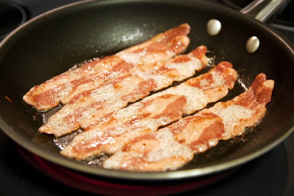 Frontal diagonal view of cooking slices turkey bacon in a fa dark colored frying pan. Dark surface in background.