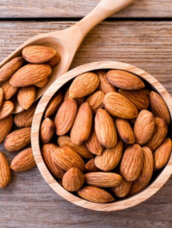 Up close aerial view of a small wooden bowl of whole almonds and a natural wood scoop overflowing with almonds onto a medium colored wood surface in the background.