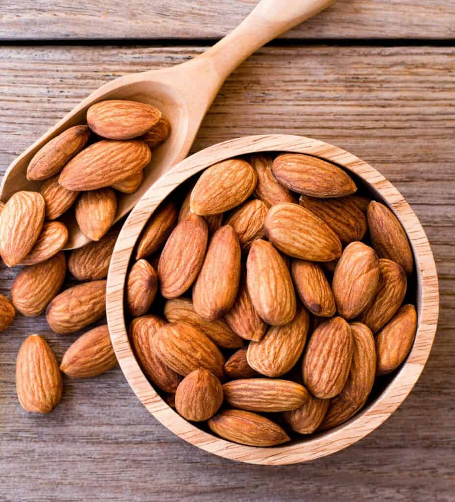 Up close aerial view of a small wooden bowl of whole almonds and a natural wood scoop overflowing with almonds onto a medium colored wood surface in the background.