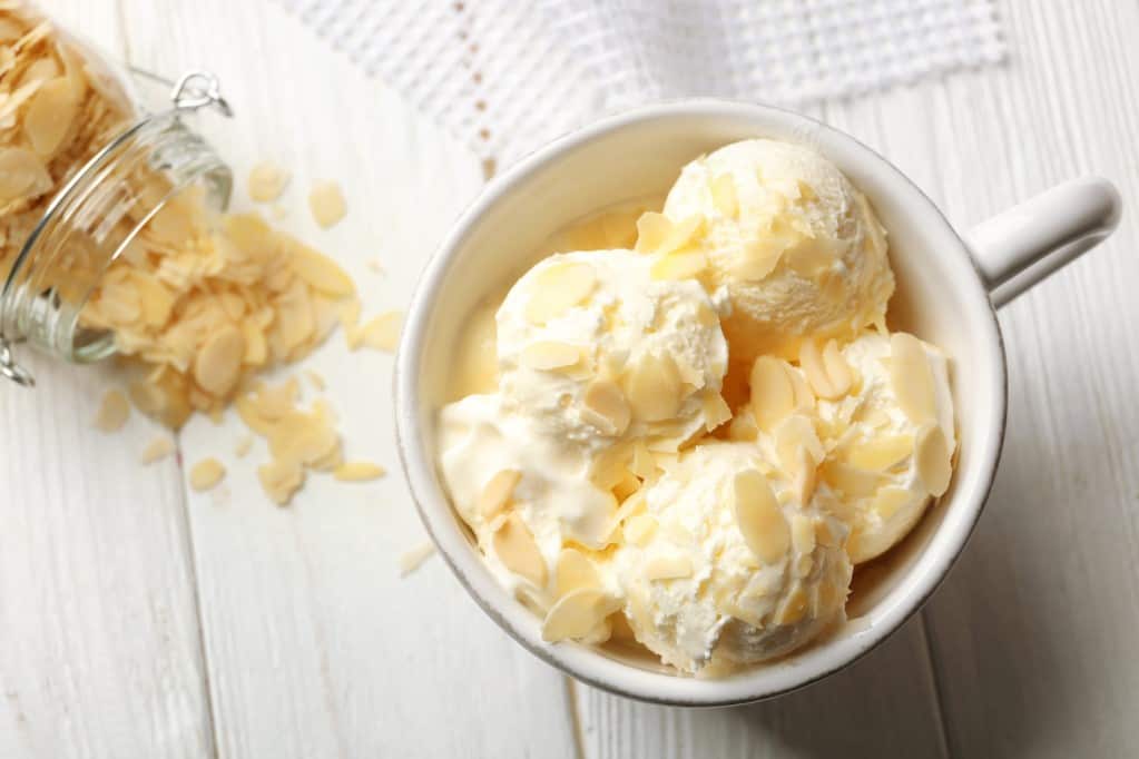 Aerial view of a white ceramic mug filled with four small scoops of vanilla ice cream topped with sliced almonds and a clear glass jar tipped over spilling almond slices spilling onto a whitewashed wooden background.