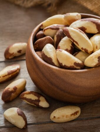 Diagonal shot of a small wooden bowl filled and overflowing with raw Brazil nuts with a burlap cloth in the background, all resting on a wooden surface.