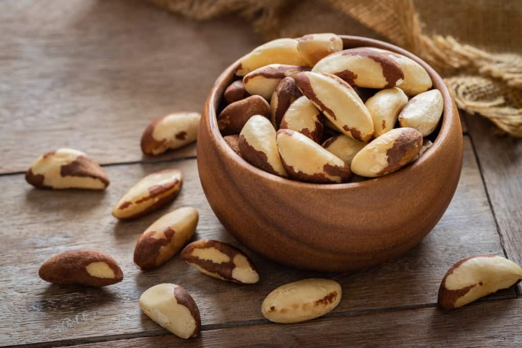 Diagonal shot of a small wooden bowl filled and overflowing with raw Brazil nuts with a burlap cloth in the background, all resting on a wooden surface.