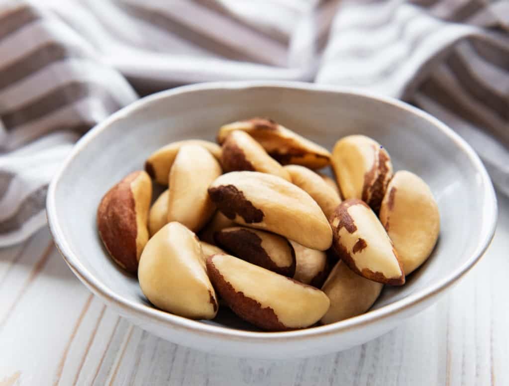 Raw Brazil nuts in a small white dish with a grey and white dishtowel in soft focus in the background, resting on a whitewashed wooden surface.